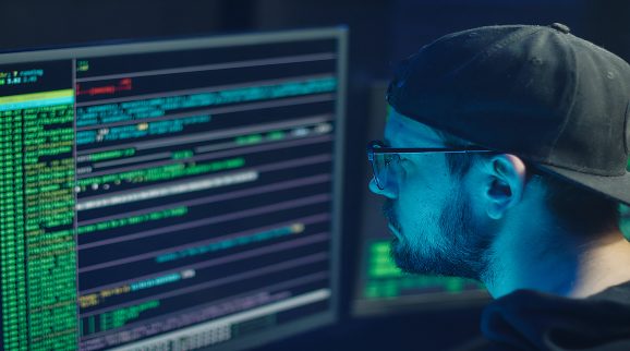 Man with a black cap and glasses looking at a computer screen with code on it - assumed to be a scammer or involved in some type of fraud