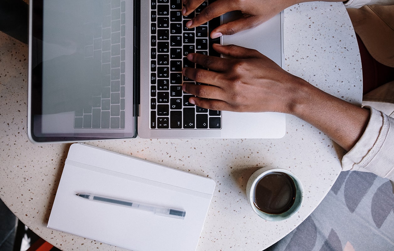 close up shot of someone typing on their macbook, on the table next to it is a cup of coffee, pen and notebook